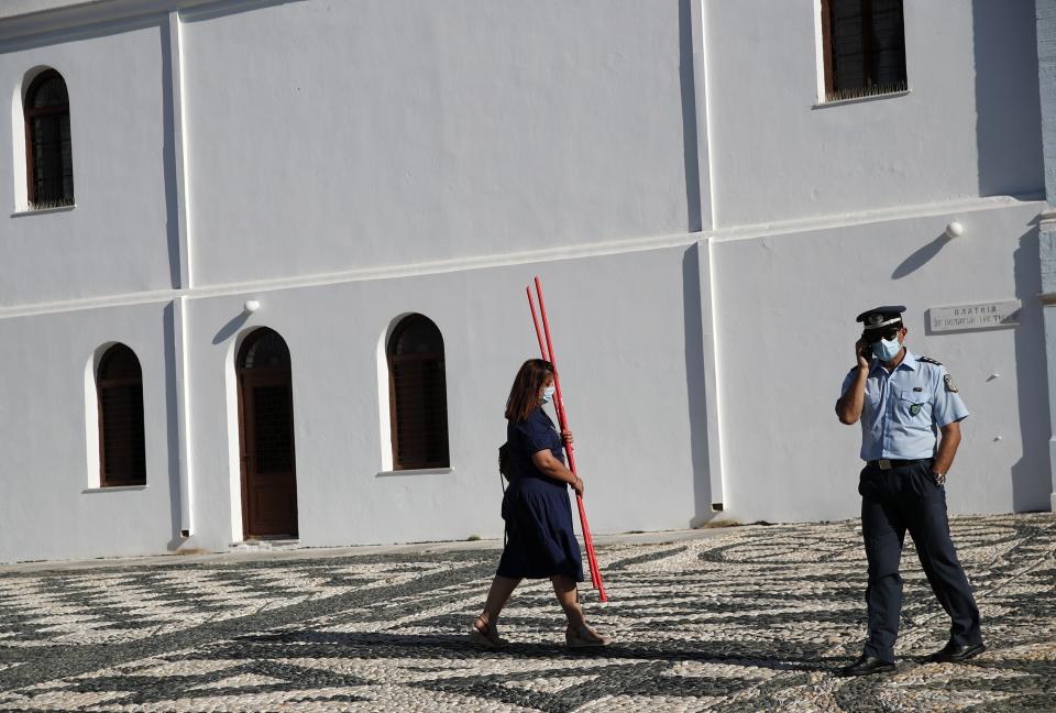 A woman wearing a face mask against the spread of the new coronavirus, holds candles as a policeman speaks on his cellphone outside the Holy Church of Panagia, on the Aegean island of Tinos, Greece, on Saturday, Aug. 15, 2020. For nearly 200 years, Greek Orthodox faithful have flocked to Tinos for the August 15 feast day of the Assumption of the Virgin Mary, the most revered religious holiday in the Orthodox calendar after Easter. But this year there was no procession, the ceremony _ like so many lives across the globe _ upended by the coronavirus pandemic. (AP Photo/Thanassis Stavrakis)