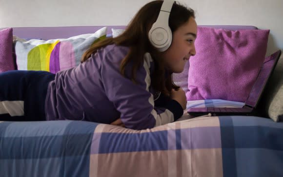 Girl lying on daybed smiling, wearing white headphones while watching something on laptop.