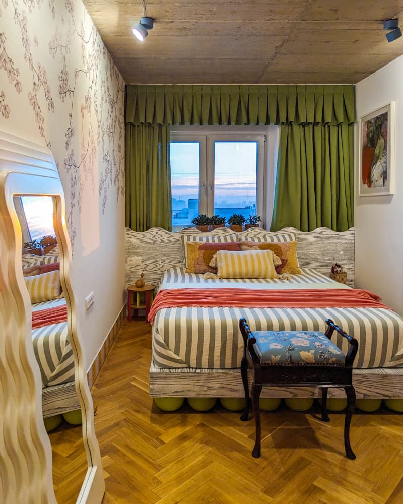 Bedroom with tree branch wallpaper wall, green curtains, parquet wood floor, and pops of stripes, yellow and orange