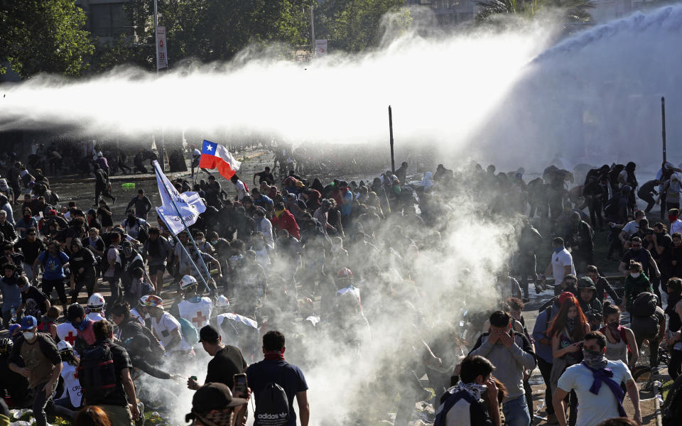 Demonstrators run from police launching water canons and tear gas as a state of emergency remains in effect in Santiago, Chile, Sunday, Oct. 20, 2019. Protests in the country have spilled over into a new day, even after President Sebastian Pinera cancelled the subway fare hike that prompted massive and violent demonstrations. (AP Photo/Esteban Felix)