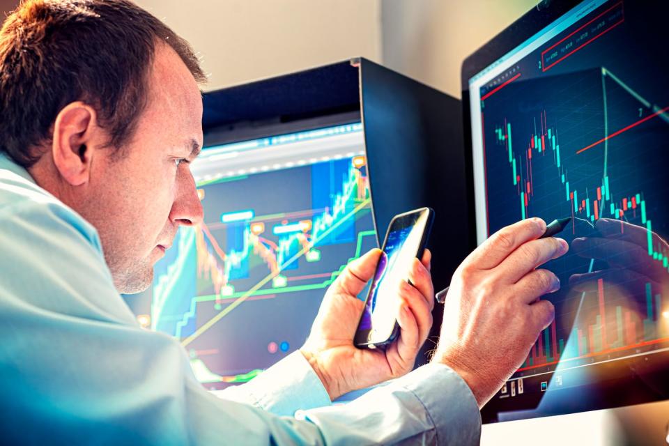 Seorang pengelola uang menggunakan ponsel pintar dan pena untuk menganalisis grafik saham yang ditampilkan di layar komputer.
