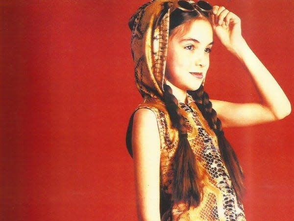 Photo of India Kushner looking off to the viewer's right against a red background. She wears a brown snakeskin dress with a hood up and sunglasses on her head. She has brown hair in braids. 