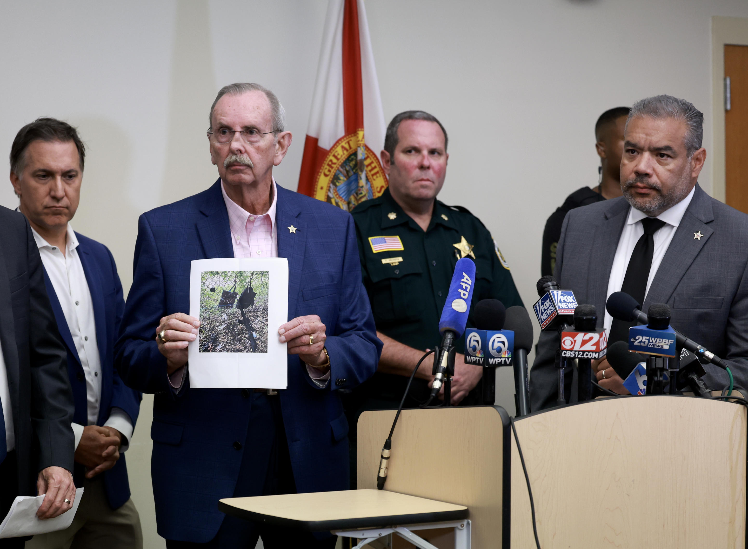 Palm Beach County Sheriff Ric Bradshaw, at a news conference, holds a photograph of the rifle and other items found near where a suspect was discovered.