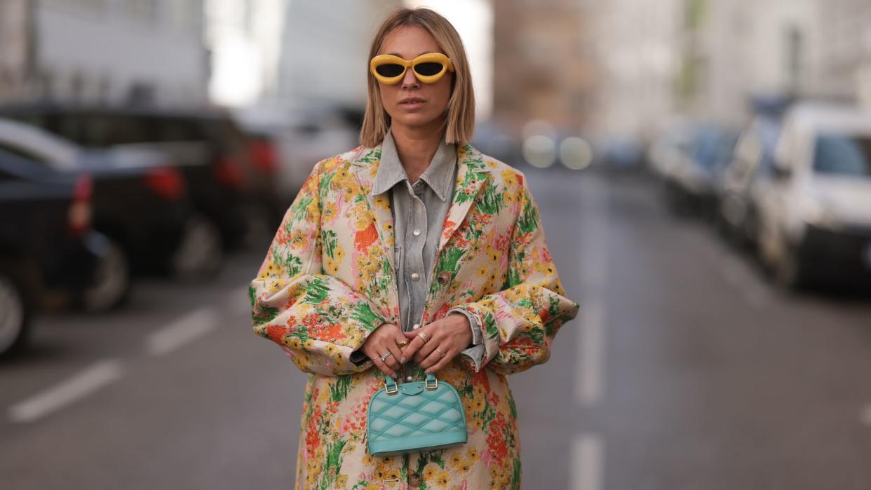 Karin Teigl seen wearing Loewe yellow sunglasses, Agolde light grey denim buttoned jeans shirt, Stine Goya yellow with colorful flower embroidered pattern long coat, Louis Vuitton light blue leather Alma mini bag, on March 23, 2024 in Munich, Germany. 