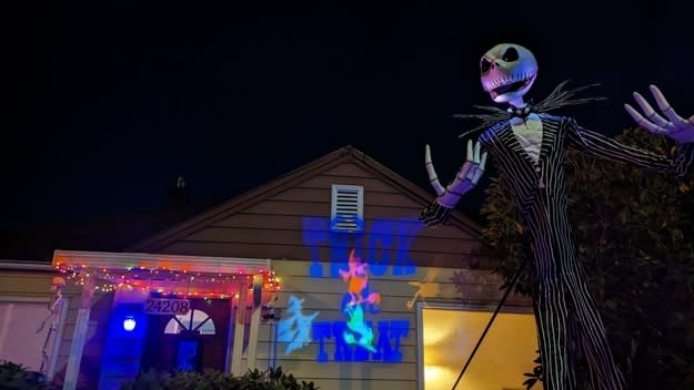 A skeleton in a pin-striped suit in front of a house at night with a "Trick or Treat" message reflecting on the house