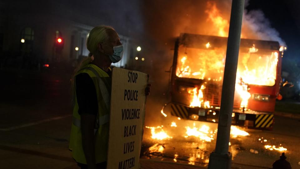 Ein Demonstrant steht mit einem Transparent vor einem brennenden Müllwagen.