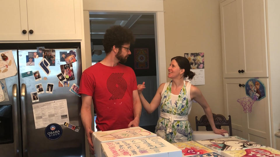 In this April 26, 2020, image from video provided by Whitney Rutz, Whitney Rutz, right, and husband, Paul, stand near several decorated boxes to hold large cinnamon rolls in their home in Portland, Ore. The Rutz family has helped raise thousands of dollars for Oregon Food Bank. (Whitney Rutz via AP)