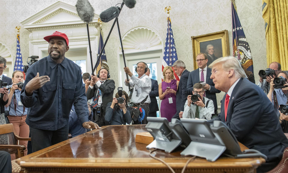 Surrounded by members of the press and others, Kanye West stands while wearing a MAGA hat with one hand on Trump's Oval Office desk while Trump sits on the other side with his elbows on the desk.