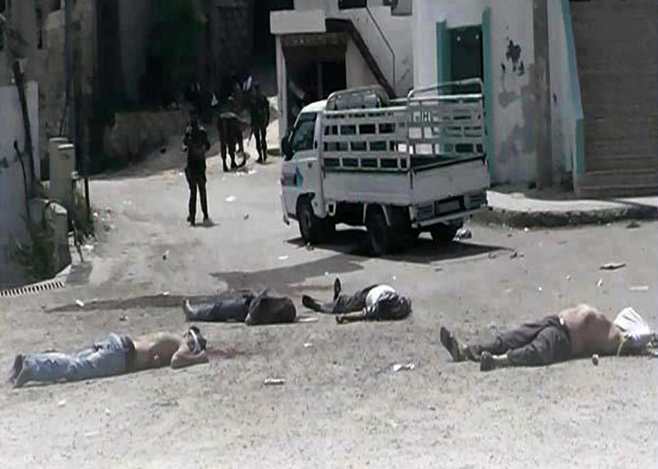 FILE - In this May 2, 2013, file photo, a citizen journalist image provided by The Syrian Revolution against Bashar Assad, an activist group, which has been authenticated based on its contents and other AP reporting, shows Syrian army soldiers loyal to Bashar Assad, seen in the background, standing in front of dead bodies at Bayda village, in the mountains outside the coastal city of Banias, Syria. The sectarian divisions burning across the rest of Syria have become physical barriers in this town on the Mediterranean coast, with soldiers manning sandbagged checkpoints dividing its Sunni Muslim southern neighborhoods from those of the Alawite and Christians in the north. Yet even after a mass killing of Sunnis in a village outside Banias in May, a tenuous truce keeps this town of 50,000 people locked in a frigid, divided peace in hopes the chaos engulfing the rest of the country will pass them by.(AP Photo/The Syrian Revolution against Bashar Assad, File)