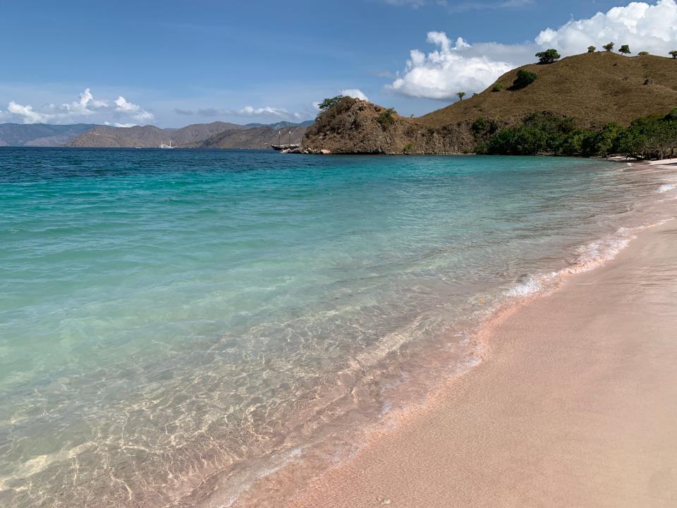 A beach with pink sand, blue water, and a lush mountainous area.