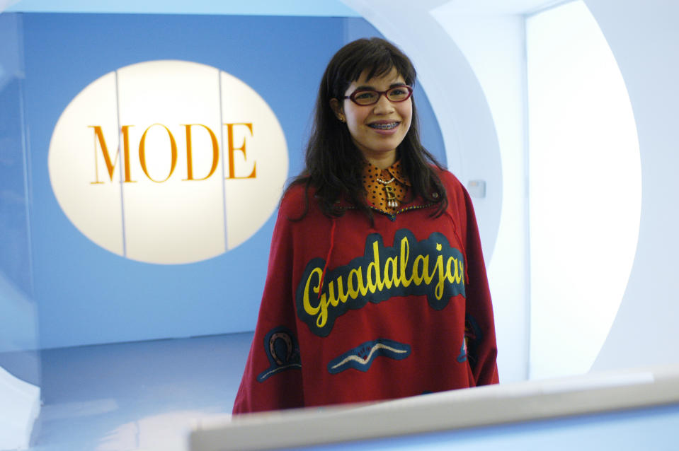 America Ferrera, as Betty Suarez, standing in front of a Mode magazine sign, wearing a red 'Guadalajara' poncho and glasses, from the TV show Ugly Betty