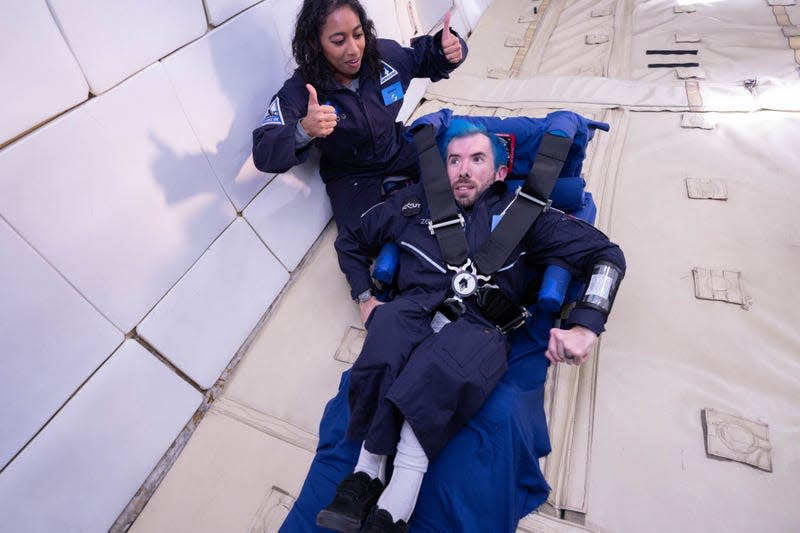 Eric Ingram, a returning flyer from AstroAccess’s inaugural 2021 mission, practices getting in and out of a 5-point harness attached to a simulated space capsule seat. He is joined by astronaut and aerospace executive Sirisha Bandla. 