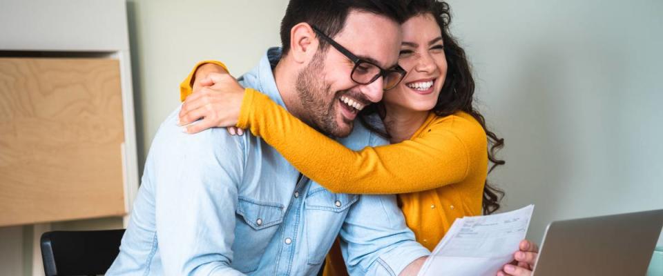 Happy couple, woman hugging man, looking at bills, sitting at desk in front of laptop