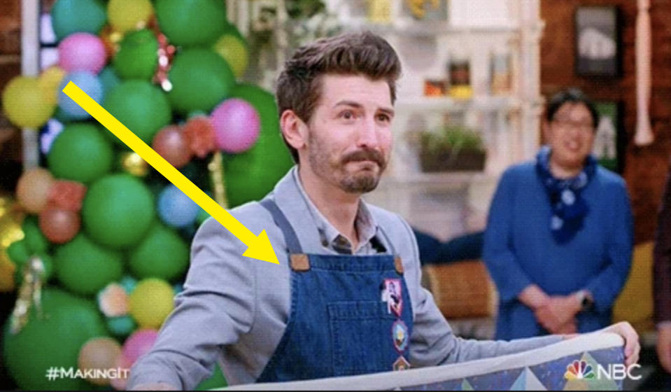 Nick Offerman on NBC's "Making It," wearing a light gray shirt and overalls, smiling. In the background, a person in blue stands near colorful decorations