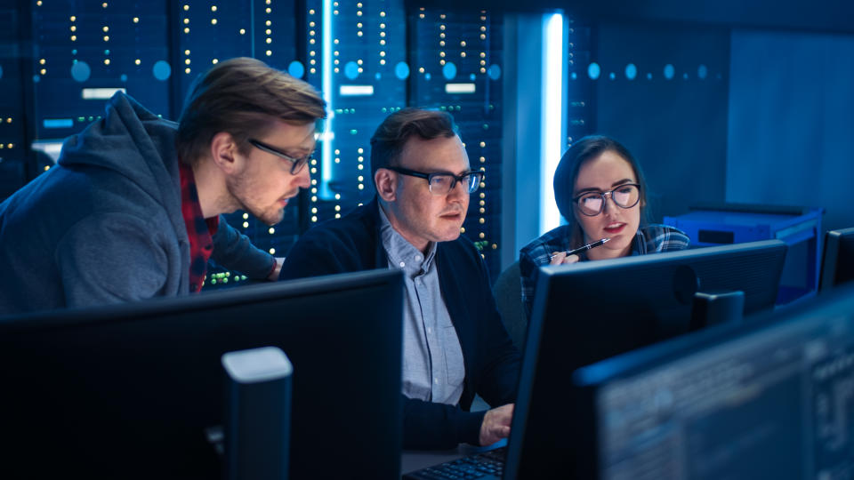 Three IT Engineers / Programmers Talking about Work, Using Computers. Technical Department with Data Server Racks. Software Development / Code Writing / Website Design / Database Architecture