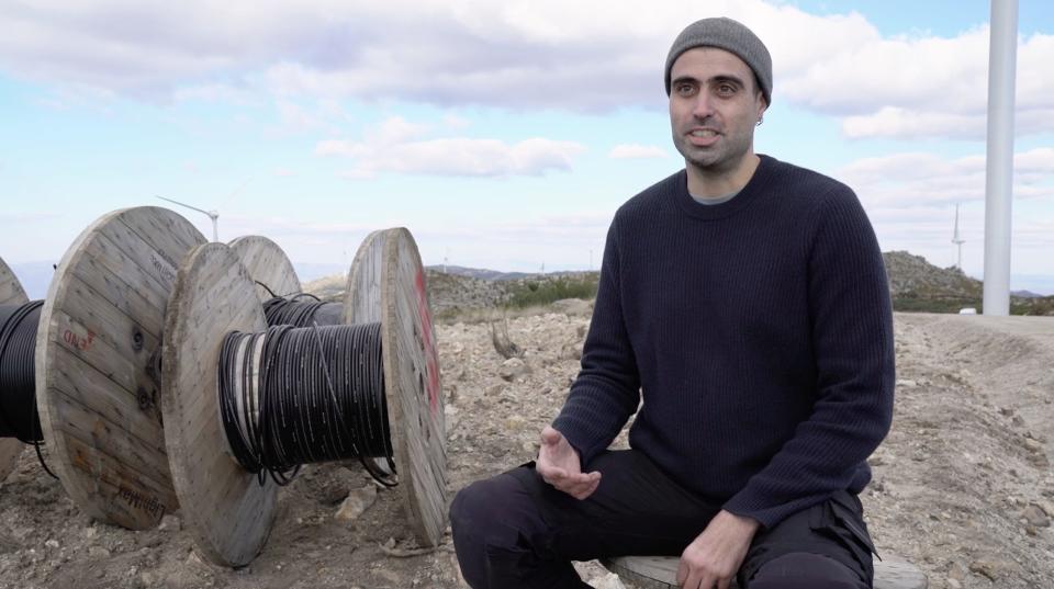 Sardo sits for an interview on a worksite with a wind turbine in the background.