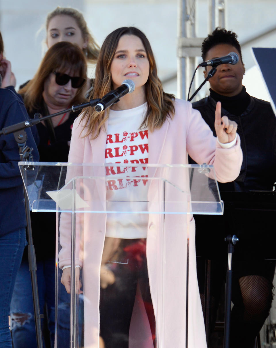 Sophia Bush at podium speaking into microphone with others in background. She wears a graphic tee and pink coat