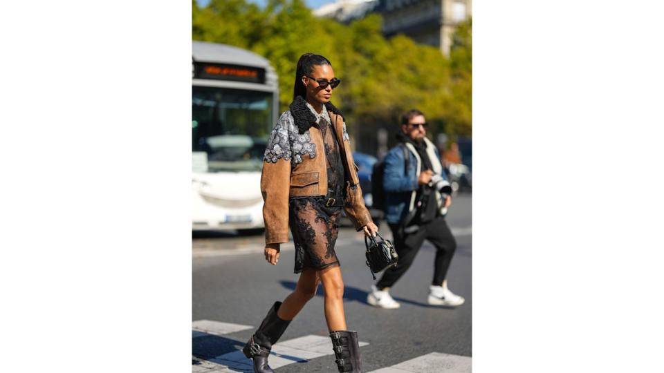 Cindy Bruna wears black sunglasses, diamonds long earrings, a black lace print pattern with diamonds embroidered collar shirt short dress , a brown shiny leather with embroidered crystal shoulder jewels jacket from Miu Miu, a black shiny leather belt, a black embossed stripped pattern handbag from Miu Miu, a lack shiny leather belted knees boots / high boot, outside Miu Miu, during Paris Fashion Week - Womenswear Spring/Summer 2023, on October 04, 2022 in Paris, France. (Photo by Edward Berthelot/Getty Images)