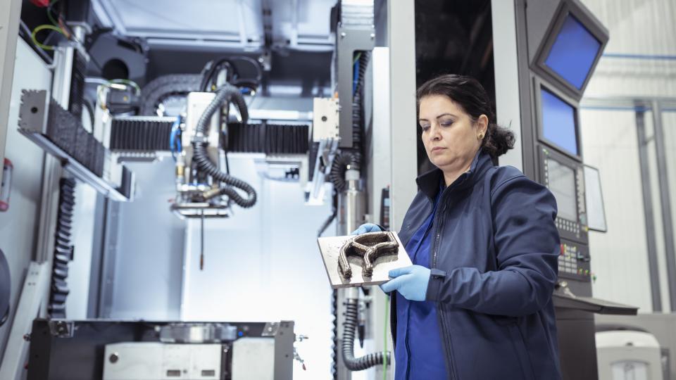 A female operator inspects a steel piece in front of a 3D printer.