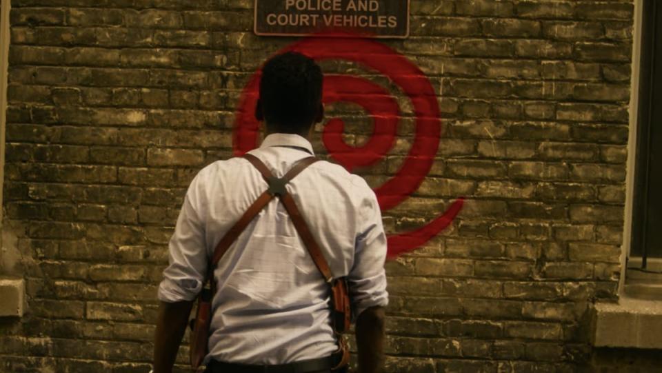 Chris Rock stands in front of a brick wall with a spiral pattern spray painted on in his Saw reboot