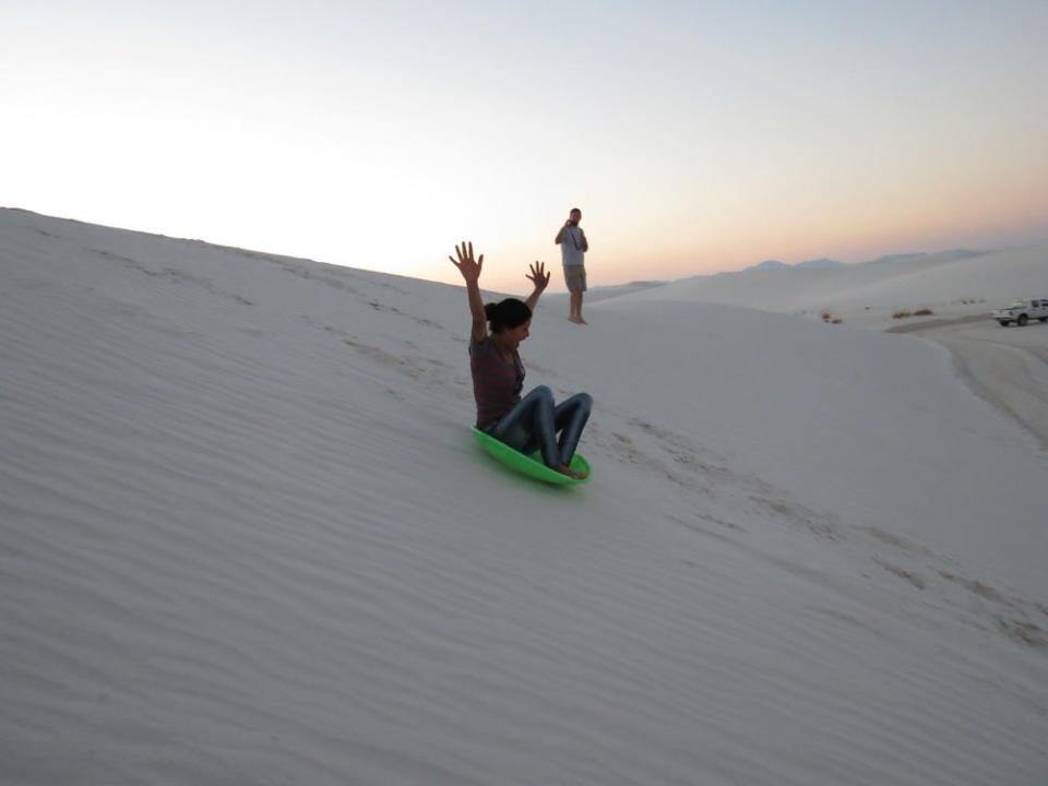 A visitor sleds down a sand dune with their hands in the air.