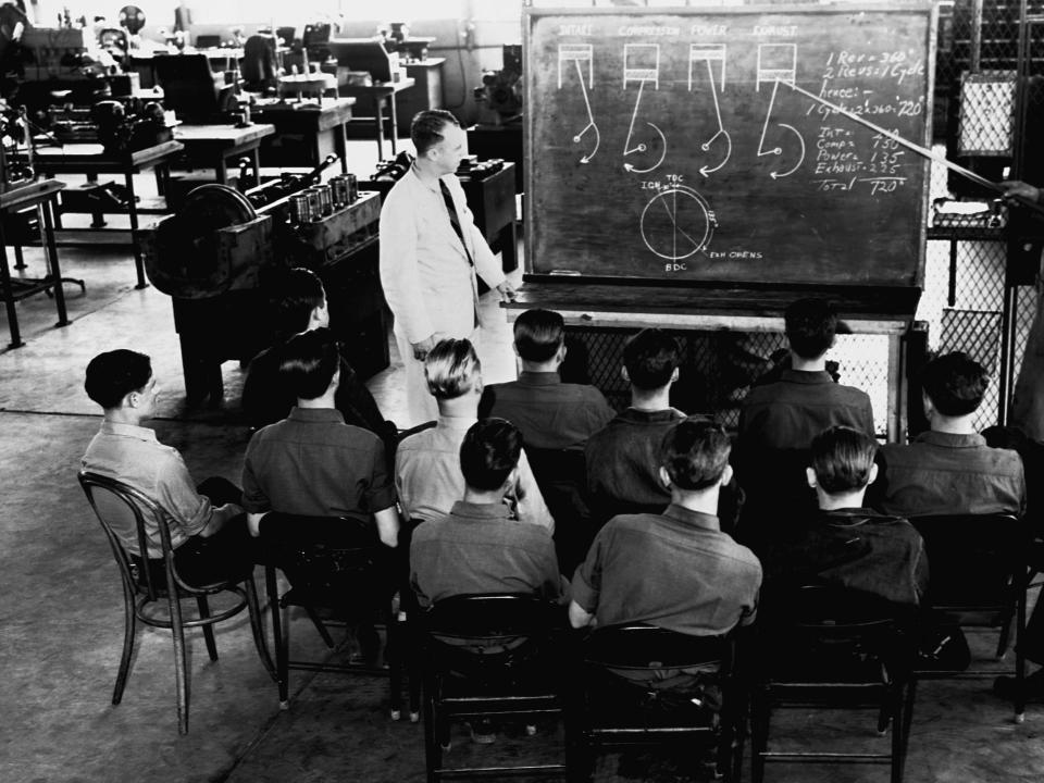A black and white photo of a man standing in front of a chalkboard giving a lesson on mechanics to a group of young men.