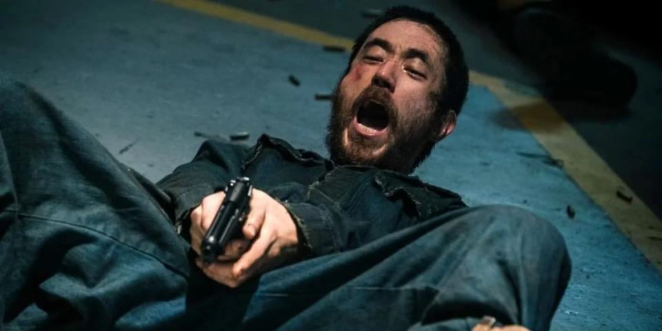 A man with trim hair and an unkempt beard lays on the floor and screams while firing a gun between his legs
