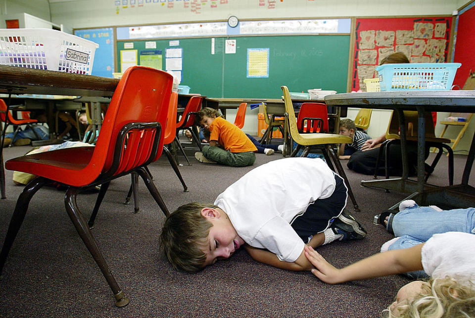 Children practicing a safety drill under desks in a classroom, lying down and covering their heads