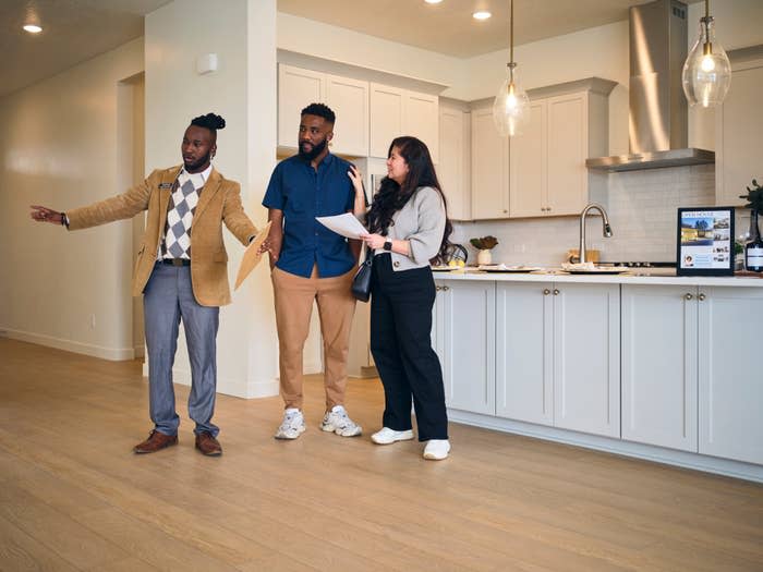 Realtor showing a kitchen to a couple, gesturing while they hold documents and listen