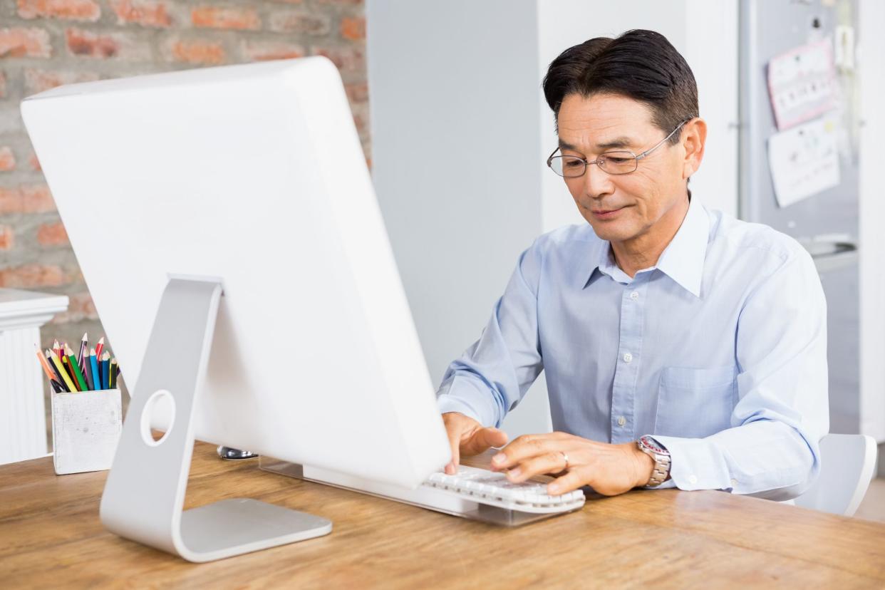 Senior man typing on a keyboard in front of a white iMac on trendy wooden desk, several colored pencils in a white holder on the desk, with both a white wall and a brick wall portion in the background