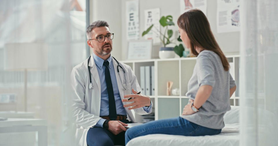 A doctor in a white coat speaks with a woman sitting on an examination table in a modern medical office