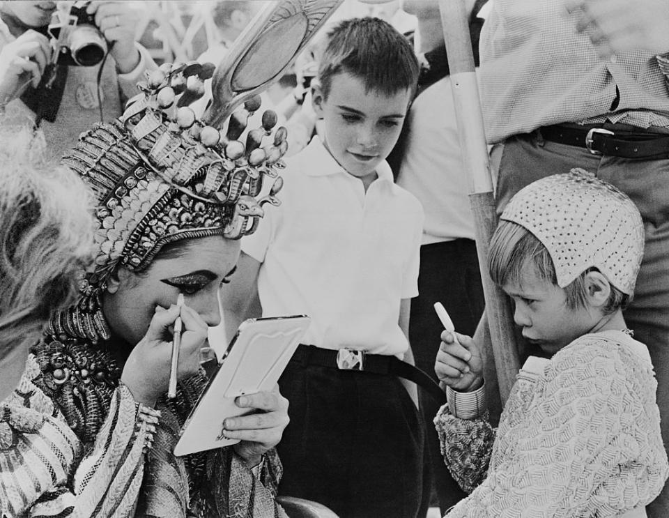 <p>Elizabeth, who did most of her dazzling makeup for the film <em>Cleopatra</em>, is seen fixing her eyeliner while her two sons look on. </p>