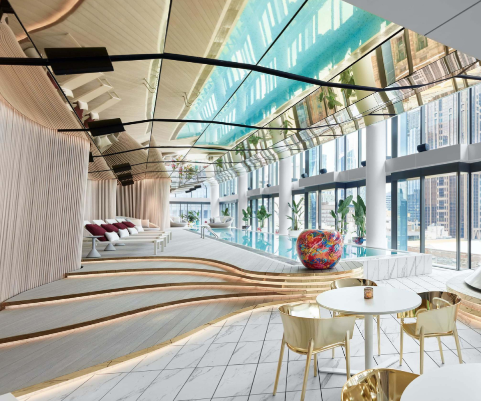 Inside W hotel's indoor pool area, with gold chairs and a table on the right foreground, a curved pale wooden staircase on the left underlit with LED lights towards the pale turquoise pool at the end with a glass wall on the right and a sloping mirror ceiling above.