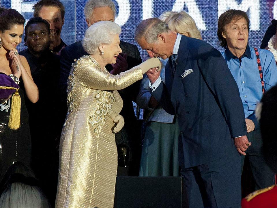 The Queen wearing a shiny gold textured long sleeve dress with gold floral and vine appliques on the front and back of the bodice. She has beige gloves on and is being kissed by Prince Charles.