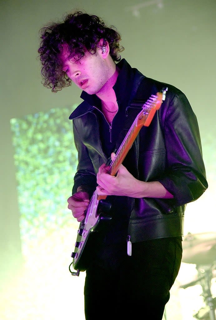 Matty in concert playing guitar, with stage lighting and screen backdrop