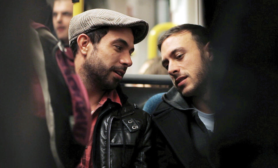 Two men are seated close to each other on a bus, engaged in a conversation. One wears a flat cap and a leather jacket, and the other wears a gray hoodie