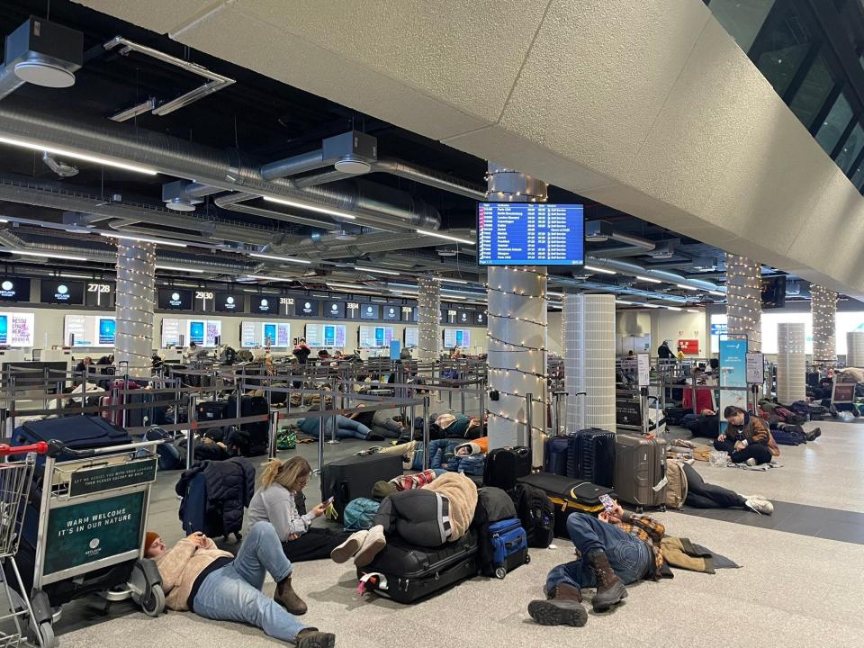 photo of stranded travelers sleeping on the ground, on top of luggage, and on the baggage claim conveyor.