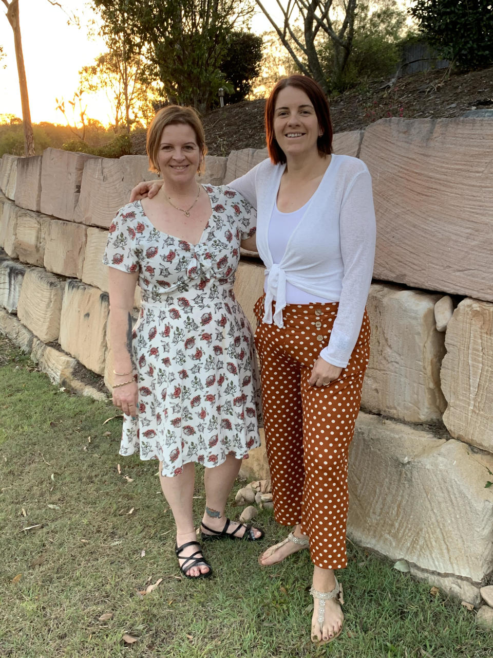 A woman wearing a printed dress poses with a woman wearing a white top and orange polka dot pants.