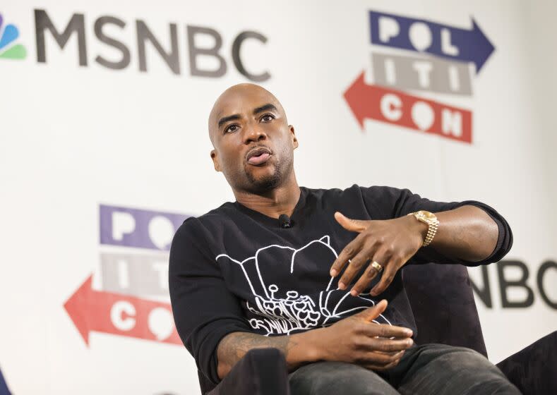 Charlamagne Tha God in a black shirt sitting down on a stage wit his hands infant of him