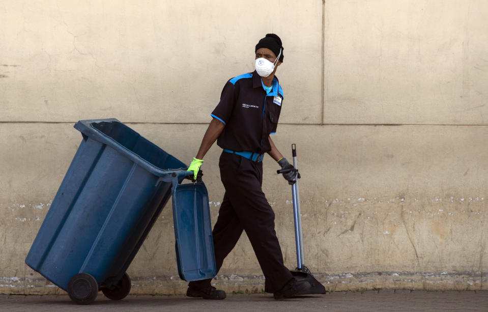 A worker wearing face masks and gloves to protect herself against coronavirus, pulls a rubbish bin whilst cleaning at Chris Hani shopping mall in Vosloorus, east of Johannesburg, South Africa, Friday, March 27, 2020. South Africa went into a nationwide lockdown for 21 days in an effort to mitigate the spread to the coronavirus. The new coronavirus causes mild or moderate symptoms for most people, but for some, especially older adults and people with existing health problems, it can cause more severe illness or death. (AP Photo/Themba Hadebe)
