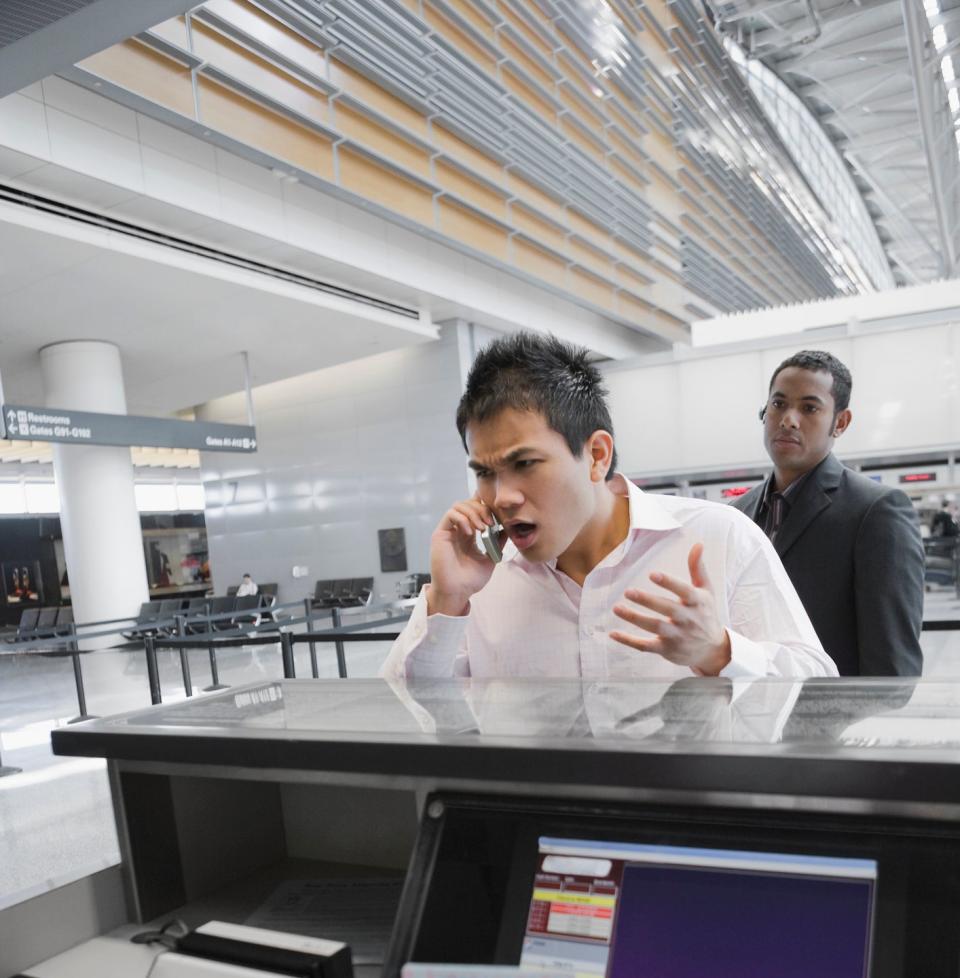 A man at an airport counter arguing on the phone while a staff member hands him a passport. Another man stands in the background, observing