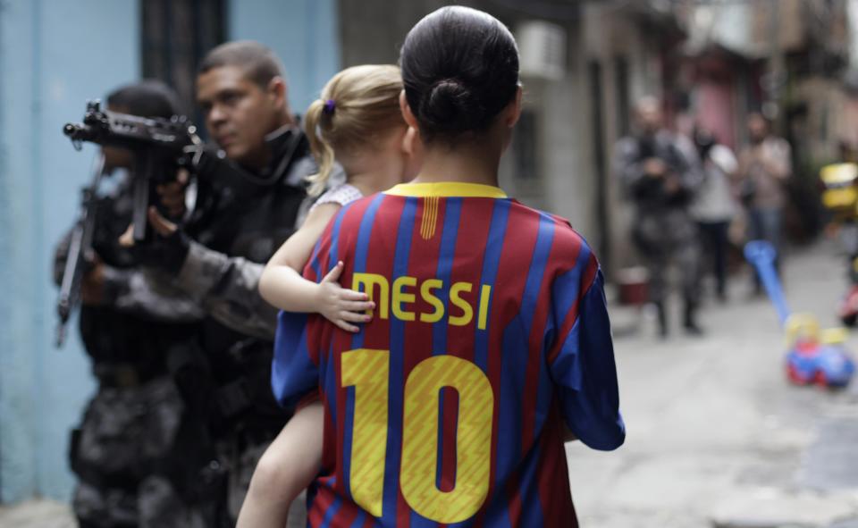 A woman wearing a Barcelona jersey with Lionel Messi's name holds a child as police officers take up positions during an operation at the Mare slums complex in Rio de Janeiro