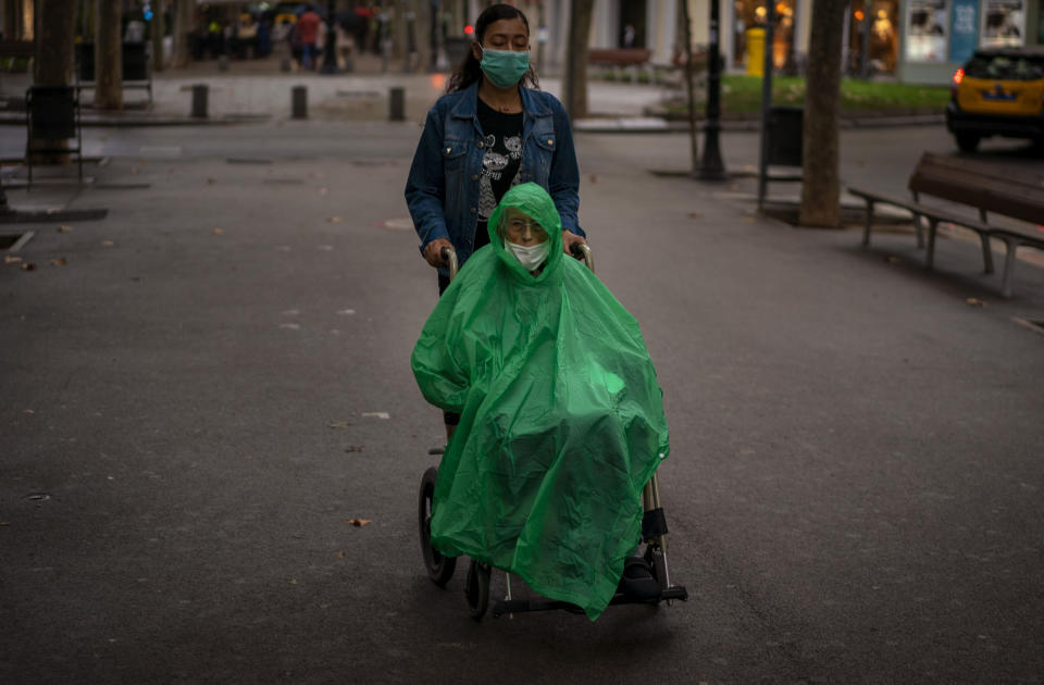 A man wearing a face mask and rain cover is pushed in a wheelchair along a boulevard in Barcelona, Spain on Wednesday Sept. 9, 2020. (AP Photo/Emilio Morenatti)