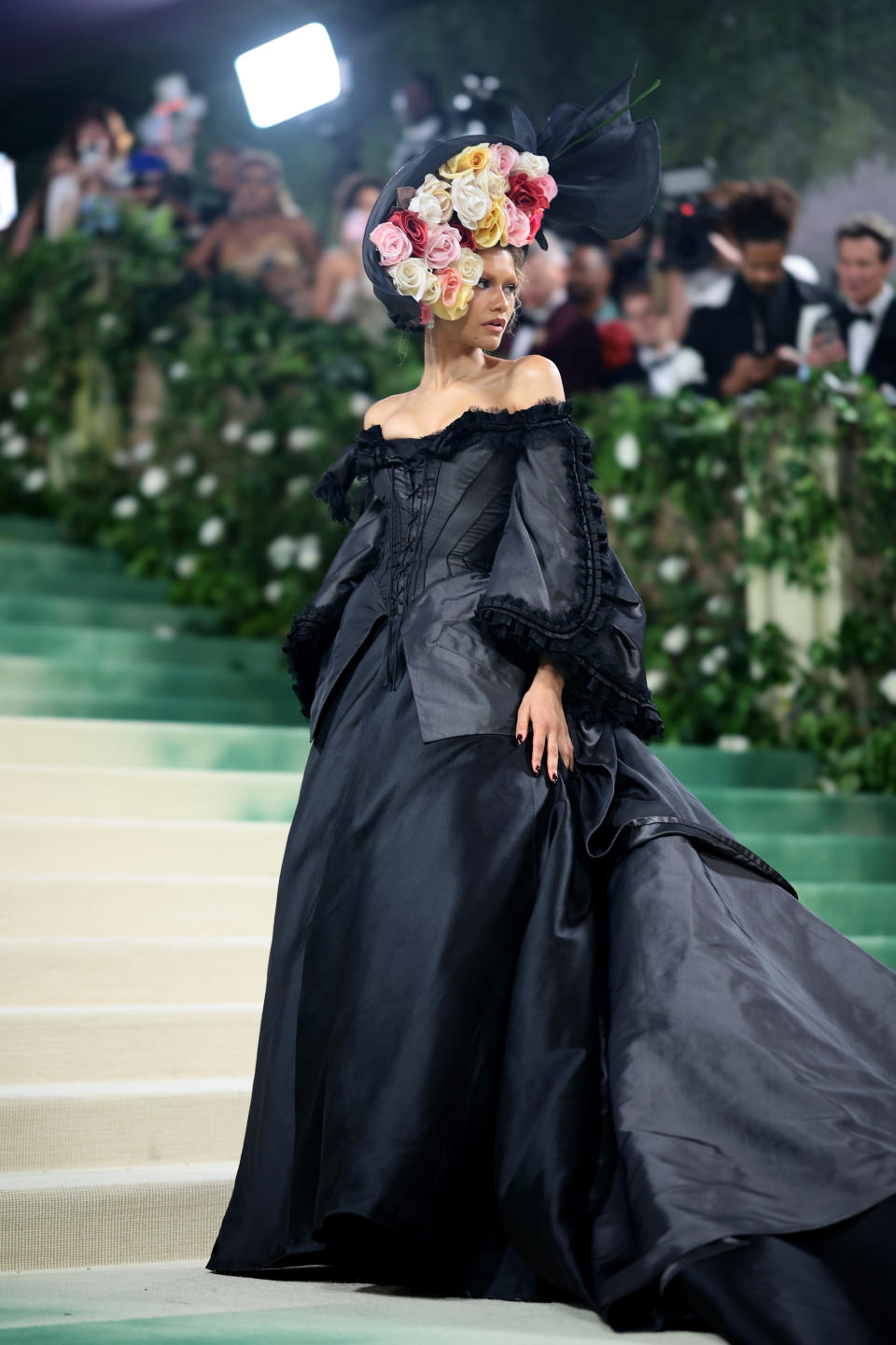 Zendaya in a dramatic off-the-shoulder black gown with an oversized floral headpiece, posing on steps