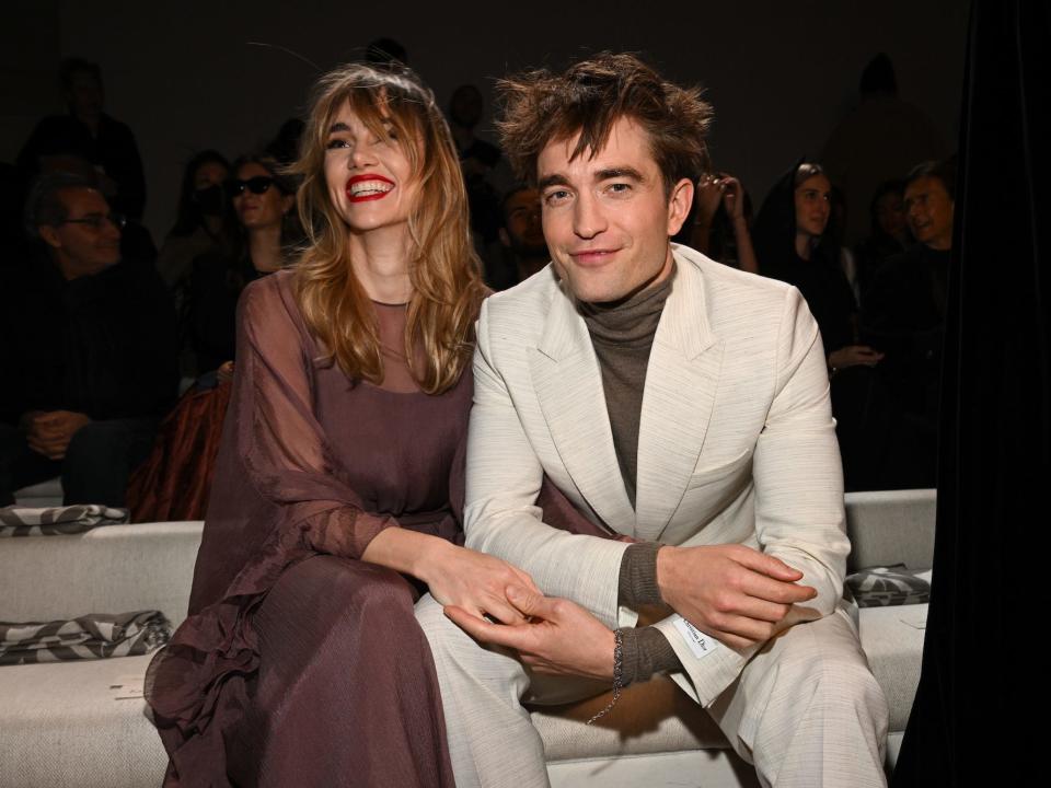 Suki Waterhouse and Robert Pattinson sit together and hold hands.