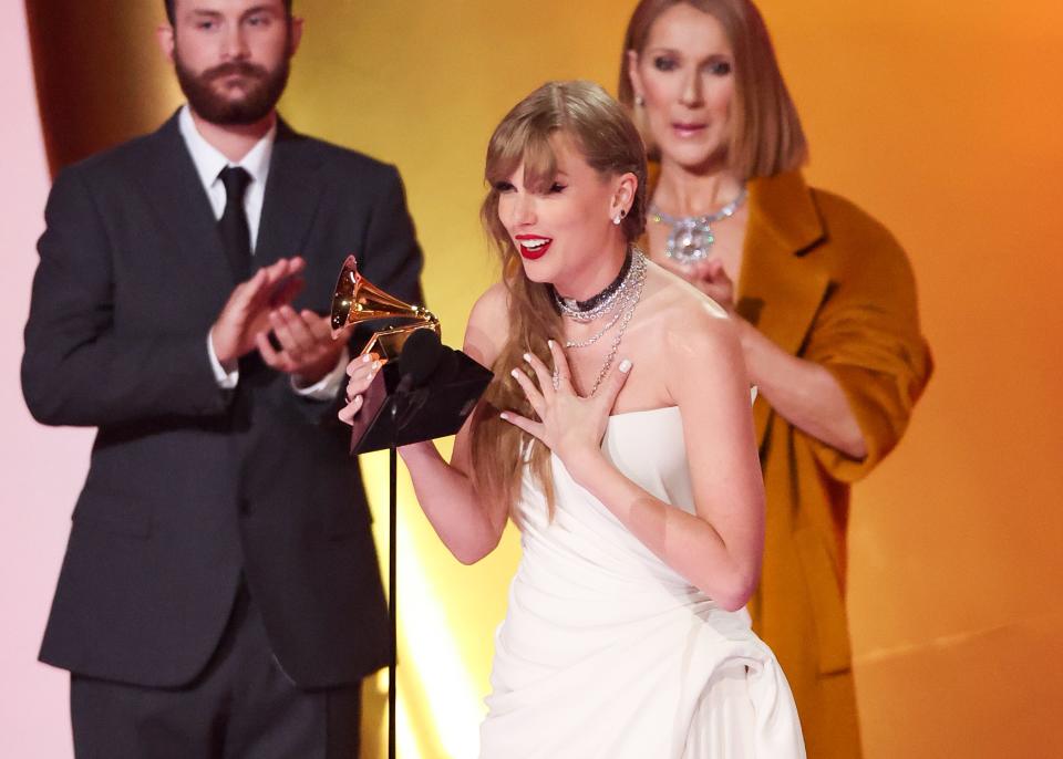 taylor swift onstage accepting a grammy award, holding the trophy in one hand and her other hand pressed to her chest. celine dion, in a brown overcoat, stands in the background