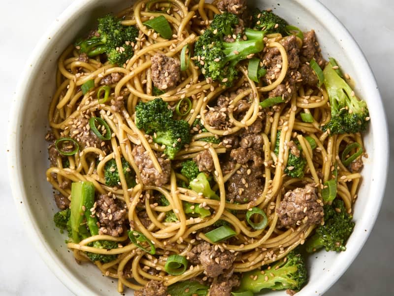 overhead shot of beef and broccoli noodles in a small white bowl, topped with sesame seeds