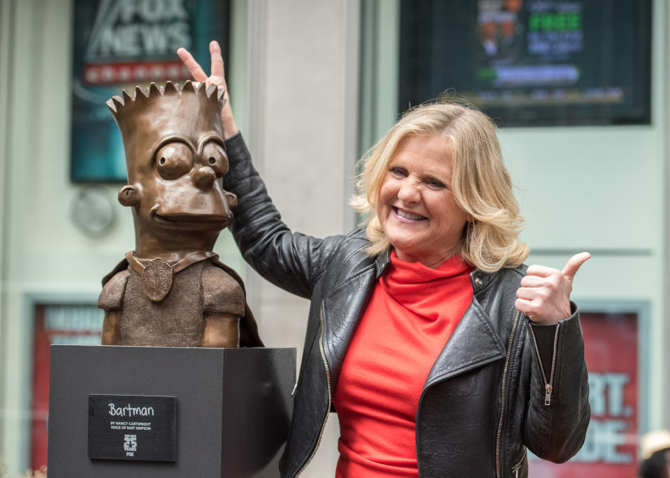 Nancy Cartwright, in a leather jacket and red top, giving a thumbs up next to a statue of Bart Simpson