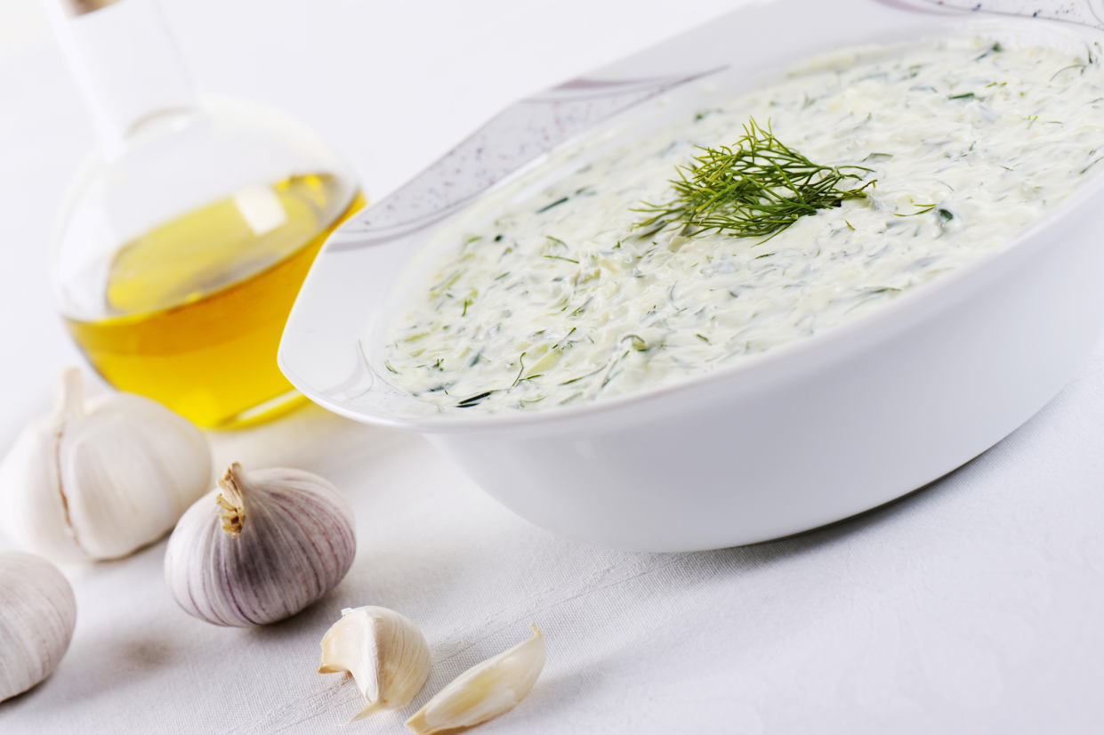 A white bowl of Tzatziki, diagonal, garlic and olive oil on the left, on a white table