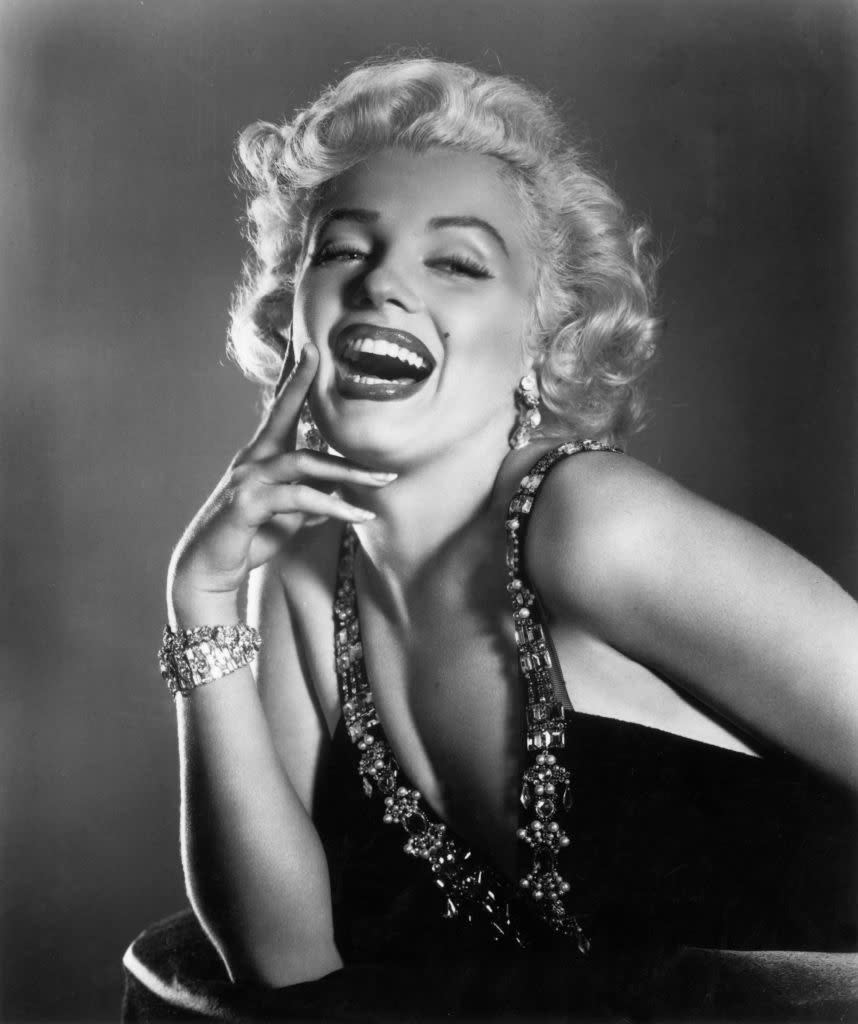 Marilyn Monroe laughing, wearing a glamorous dress with jeweled straps and a bracelet. She has styled, short curly hair